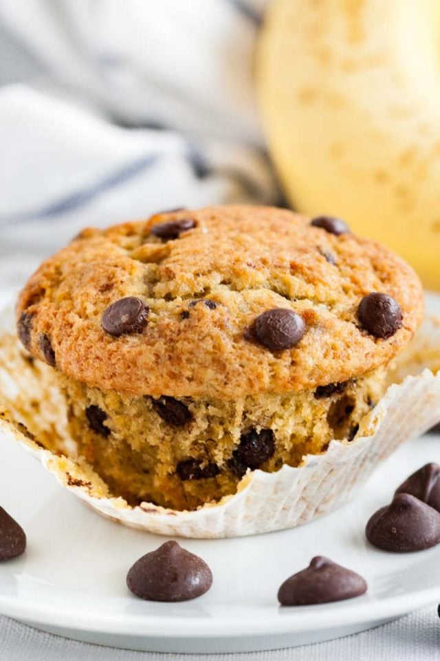 A banana chocolate chip muffin with its paper opened on a white plate garnished with chocolate chips in front of a banana.