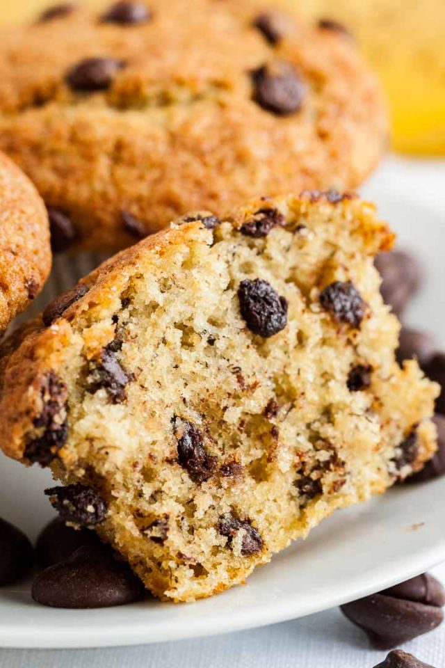 Close-up of a halved banana chocolate chip muffin on a white plate with two more muffins and some chocolate chips.
