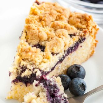 A slice of blueberry cake with streusel topping on a white plate garnished with blueberries. A fork has taken a piece out of the slice and is lying next to it. There's a glass bowl of blueberries in the background.