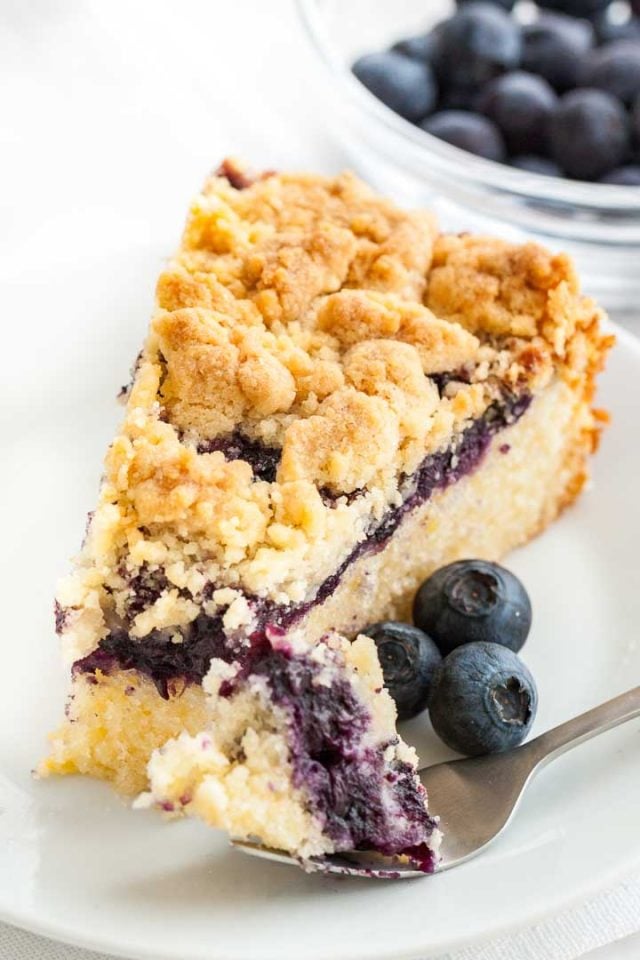 A slice of blueberry cake with streusel topping on a white plate garnished with blueberries. A fork has taken a piece out of the slice and is lying next to it. There\'s a glass bowl of blueberries in the background.