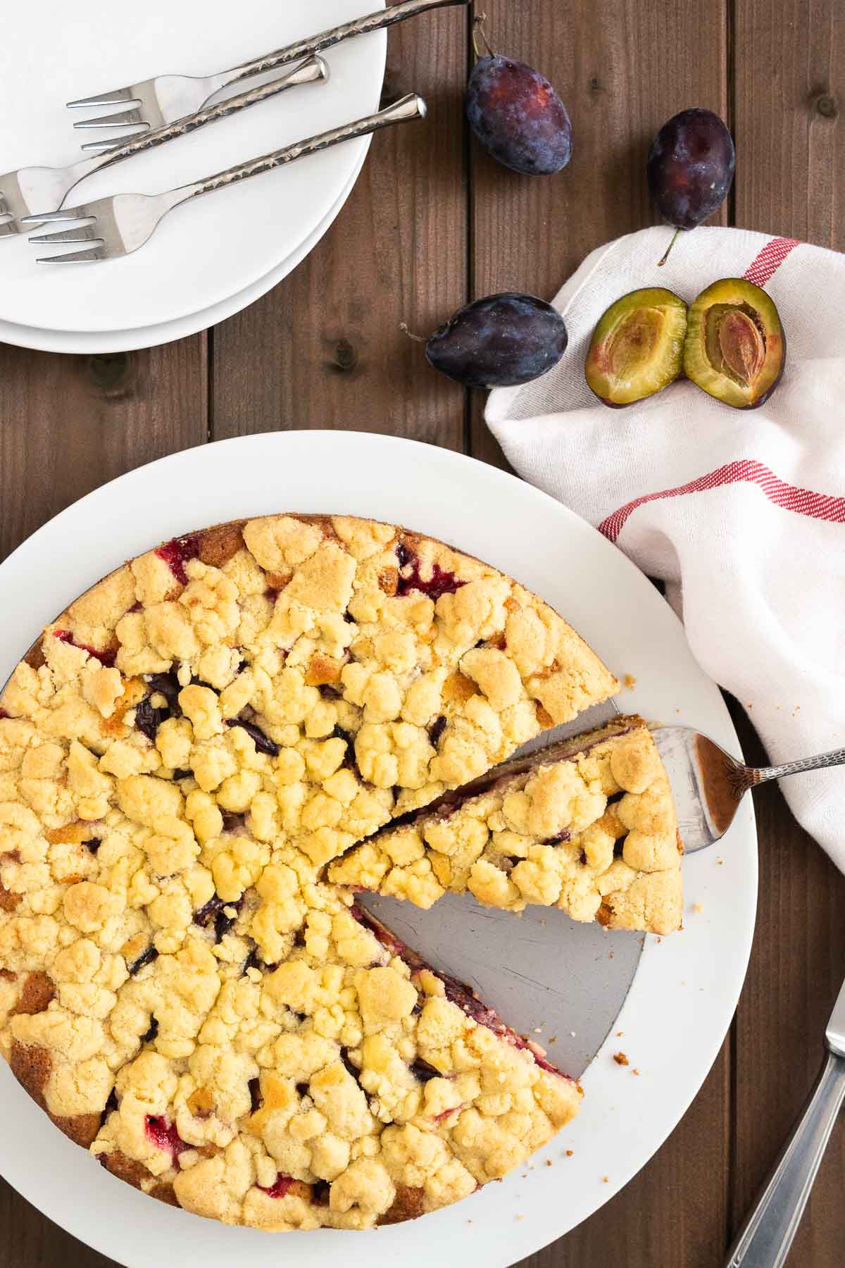 Top-down shot of a plum cake with streusel on a springform bottom on a white plate. A cake server is lifting out a slice. Next to the plate, there are plums, a knife, two white plates with forks and a white dishtowel with a red stripe.