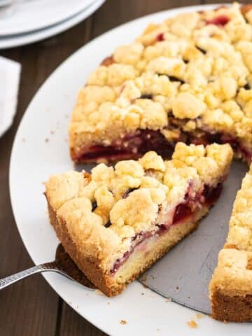 A plum cake with streusel on a springform bottom on a white plate. A cake server is lifting out a slice.
