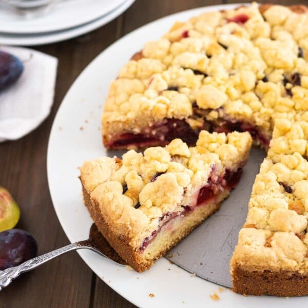 A plum cake with streusel on a springform bottom on a white plate. A cake server is lifting out a slice.