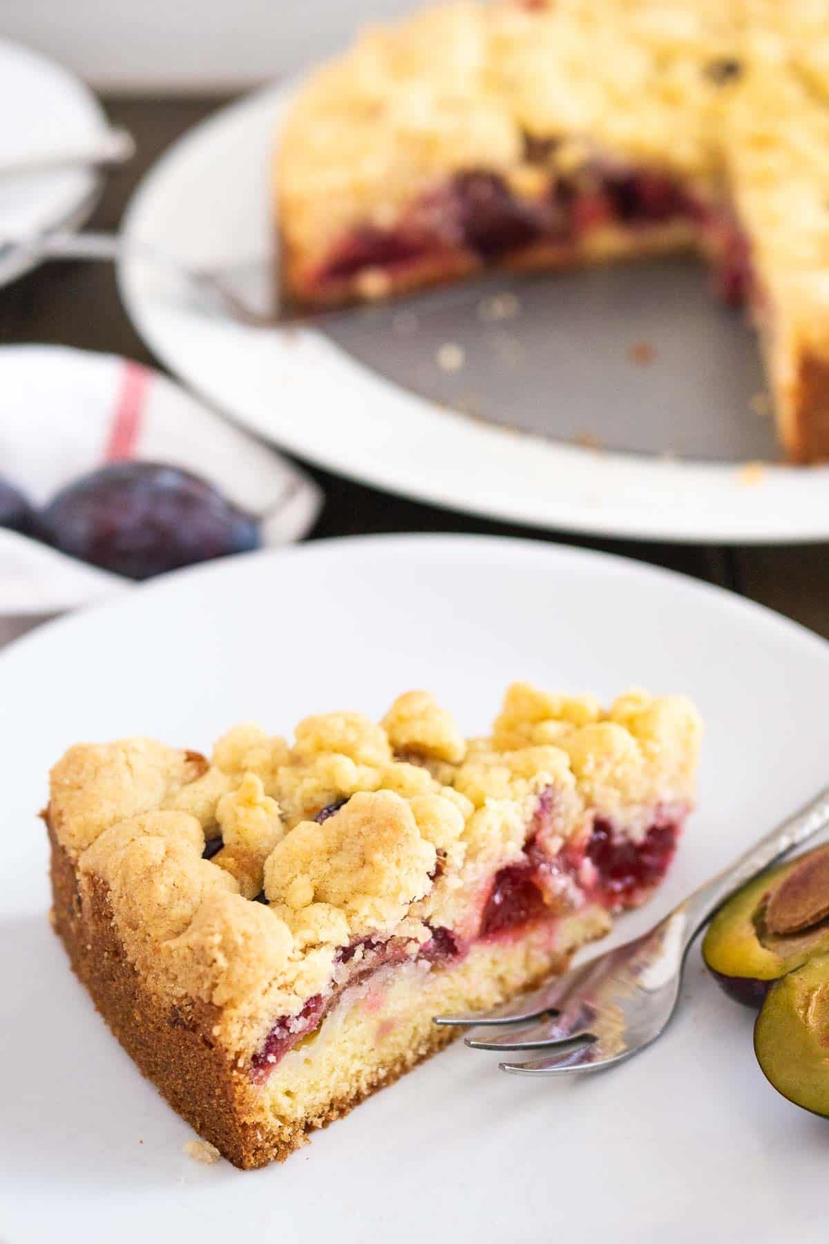 A slice of plum cake topped with streusel on a white plate with a fork and a halved plum. The whole cake is in the background.