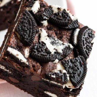 Close-up of an oreo brownie, topped with oreo cookies leaning against a white plate stacked on a pink plate.