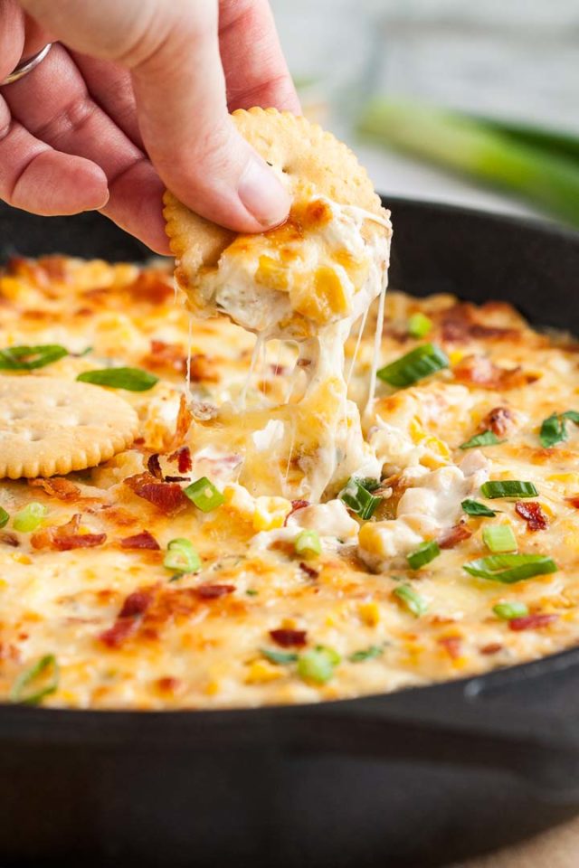 A lodge pan with corn dip and molten cheese on top. A hand is scooping out the dip with a cracker.