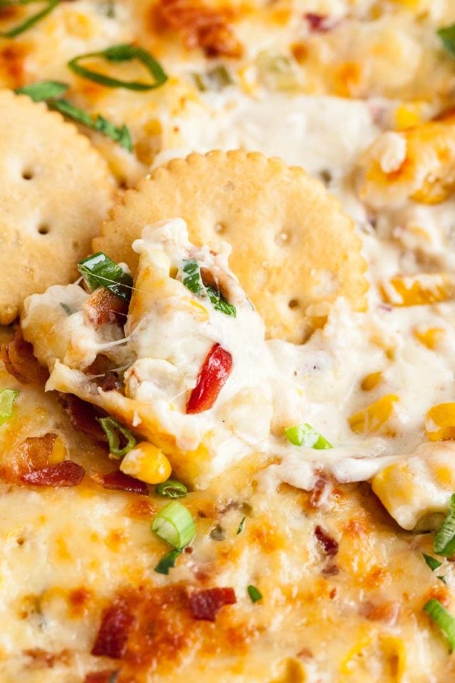 Close-up of hot corn dip, containing peppers, corn, chives and cheese with some crackers.