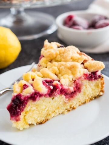 A slice of lemon raspberry cake with streusel topping on a white plate with a fork. There is a whole lemon and some frozen raspberries in the background.