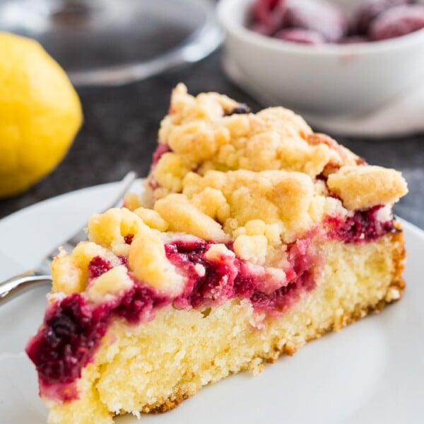 A slice of lemon raspberry cake with streusel topping on a white plate with a fork. There is a whole lemon and some frozen raspberries in the background.
