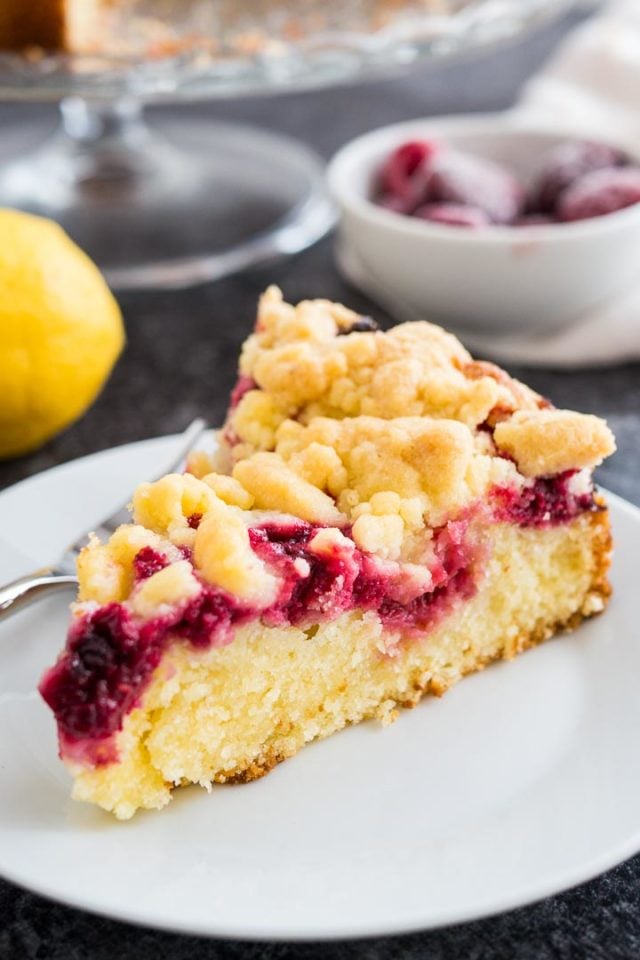 A slice of lemon raspberry cake with streusel topping on a white plate with a fork. There is a whole lemon and some frozen raspberries in the background.