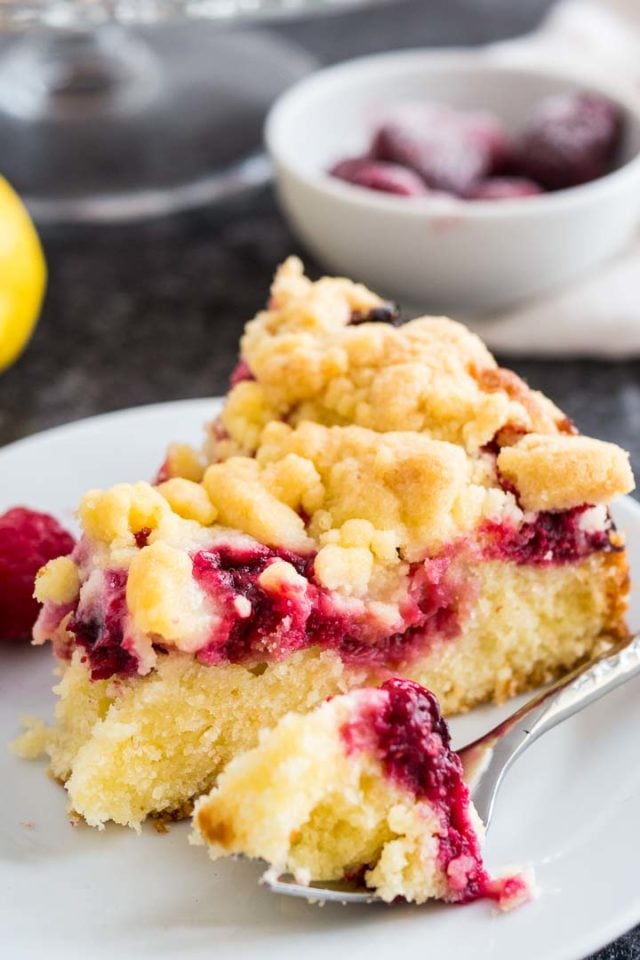 A slice of lemon raspberry cake with streusel topping on a white plate with a fork with a small piece of the cake on it, garnished with lemon and raspberries.