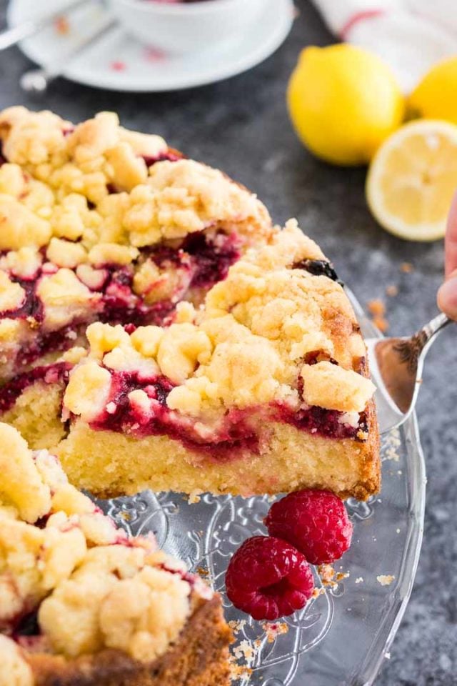 A cake server lifting a slice of lemon raspberry cake with streusel topping from a glass serving plate, garnished with lemons and raspberries.