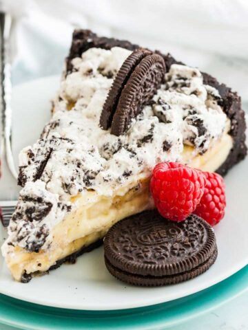 A slice of Oreo banana cream cake on a white plate with a fork, garnished with an oreo cookie and two raspberries.