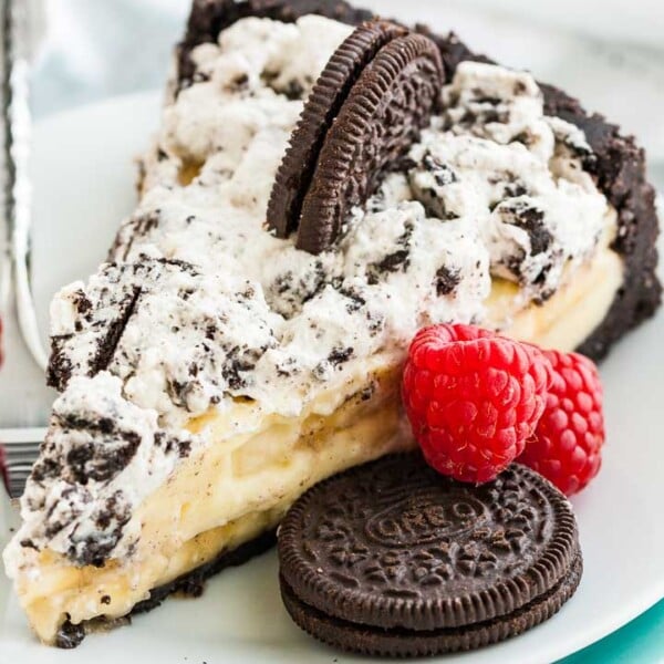 A slice of Oreo banana cream cake on a white plate with a fork, garnished with an oreo cookie and two raspberries.