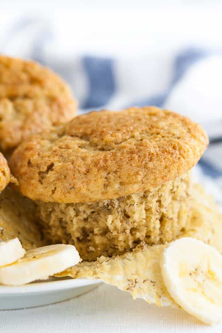 An unwrapped muffin, garnished with banana slices and a muffin and a blue and white dish towel in the background.