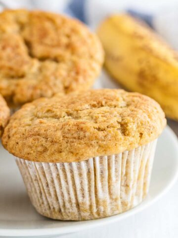 Close-up of a muffin on a white plate with several muffins and a banana in the background.