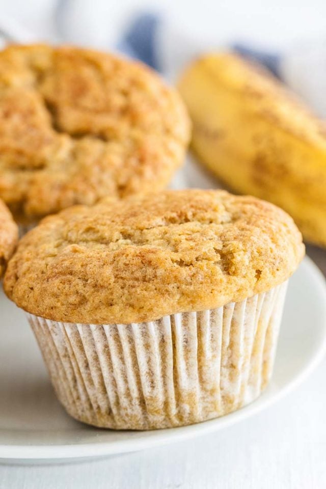 Close-up of a muffin on a white plate with several muffins and a banana in the background.