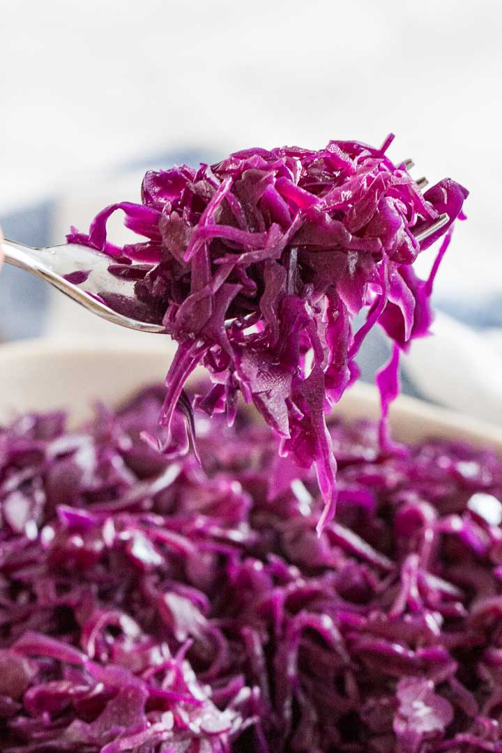 Close-up of a fork holding up some red cabbage above a white serving plate containing red cabbage.
