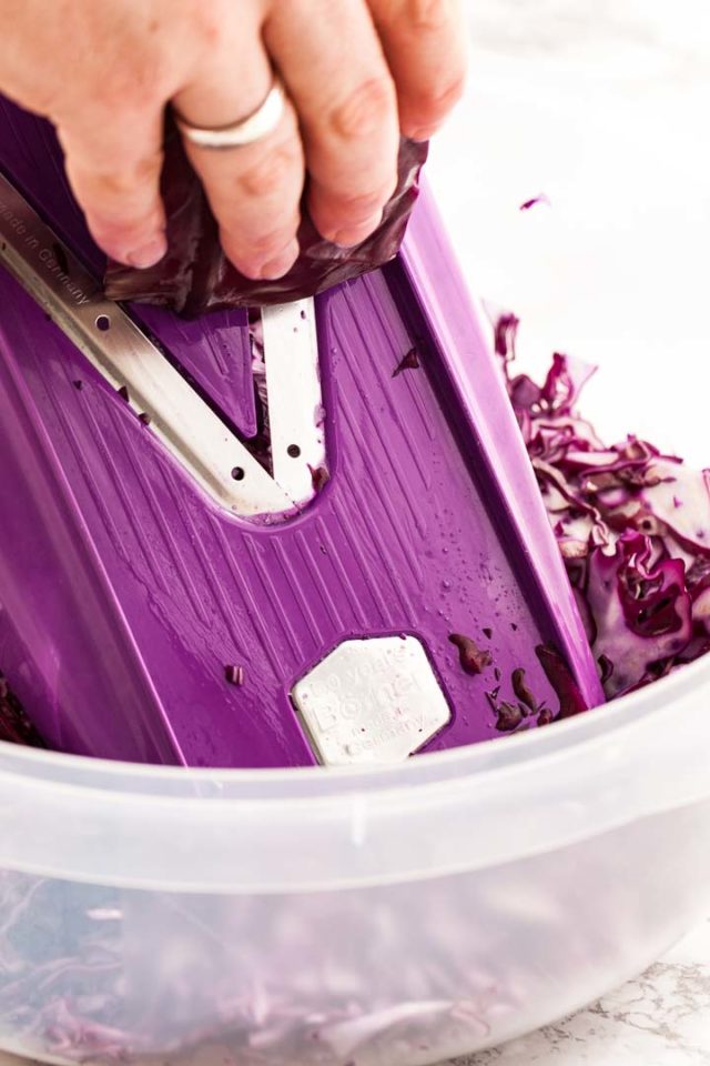 A plastic bowl with sliced, raw red cabbage and v-grater with a hand slicing red cabbage.