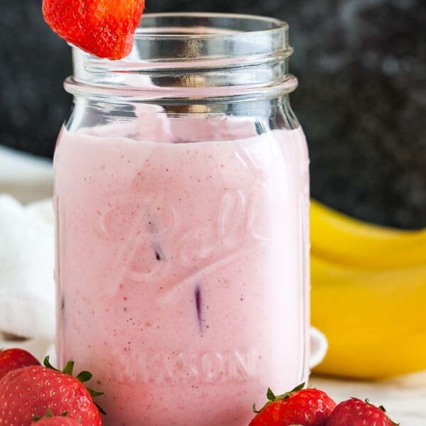 A ball jar with a strawberry banana smoothie a white straw and a strawberry on the rim on a marble surface in front of a dark background. There are some strawberries in front and 2 bananas in the back.