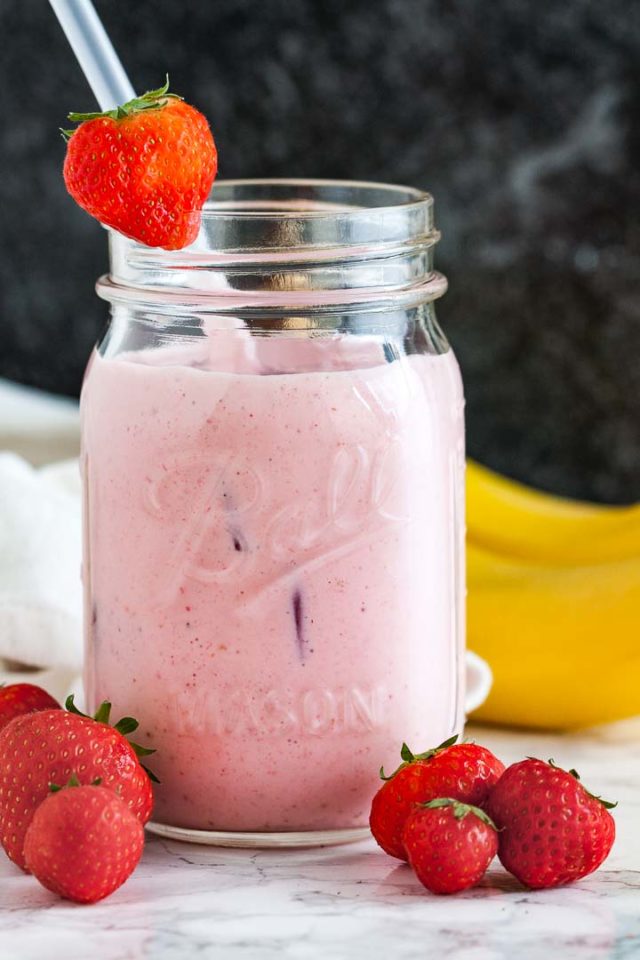 A ball jar with a strawberry banana smoothie a white straw and a strawberry on the rim on a marble surface in front of a dark background. There are some strawberries in front and 2 bananas in the back.