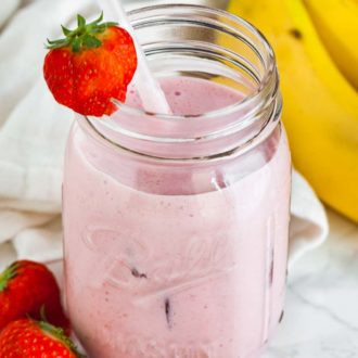 A ball jar with a strawberry banana smoothie a white straw and a strawberry on the rim on a marble surface. There's a white dish towel, 2 strawberries and some bananas next to it.