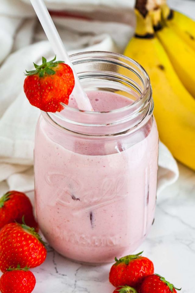 A ball jar with a strawberry banana smoothie a white straw and a strawberry on the rim on a marble surface. There\'s a white dish towel, 2 strawberries and some bananas next to it.