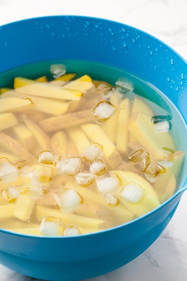 A blue bowl filled with potatoes, cut into sticks and submerged in ice water.