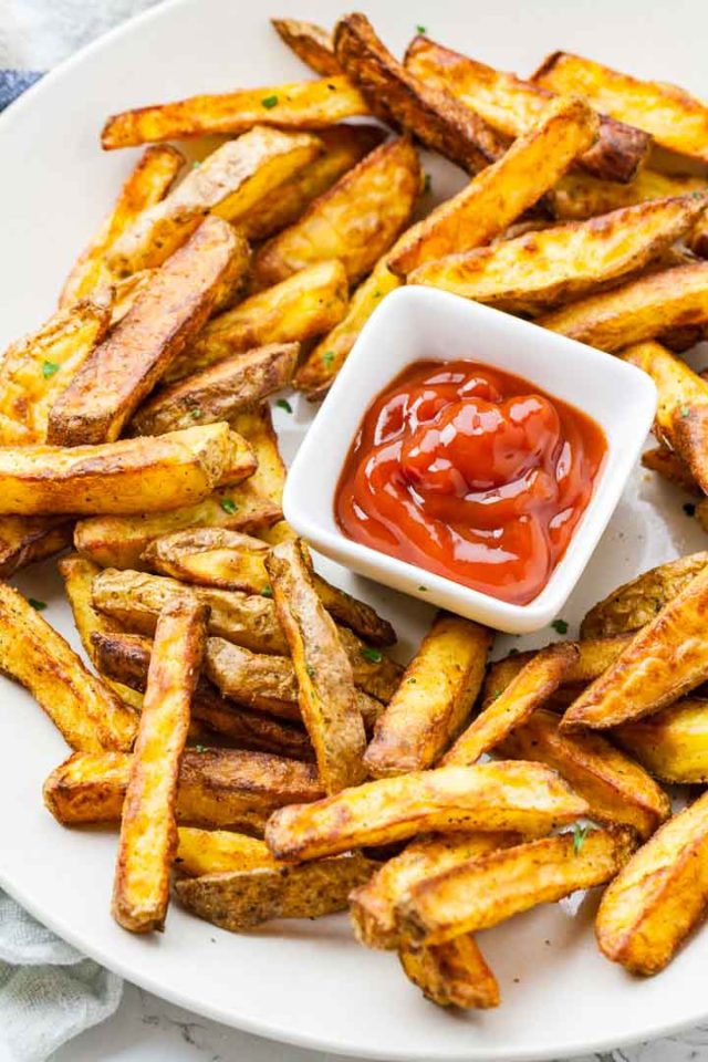 Brown, crispy fries on a white plate, with a small, square, white bowl of ketchup.