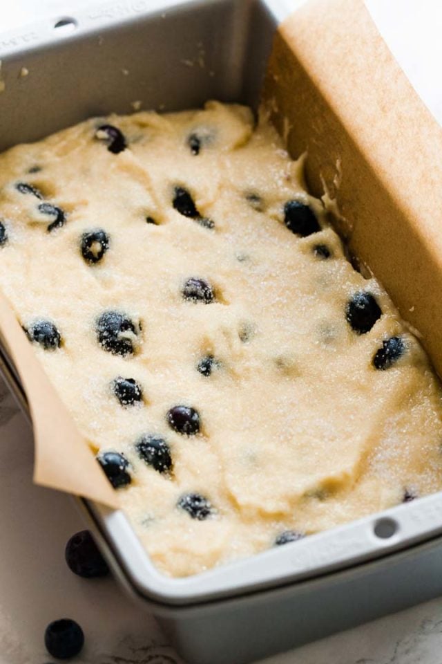A cake pan, lined with parchment paper, filled with dough with blueberries.