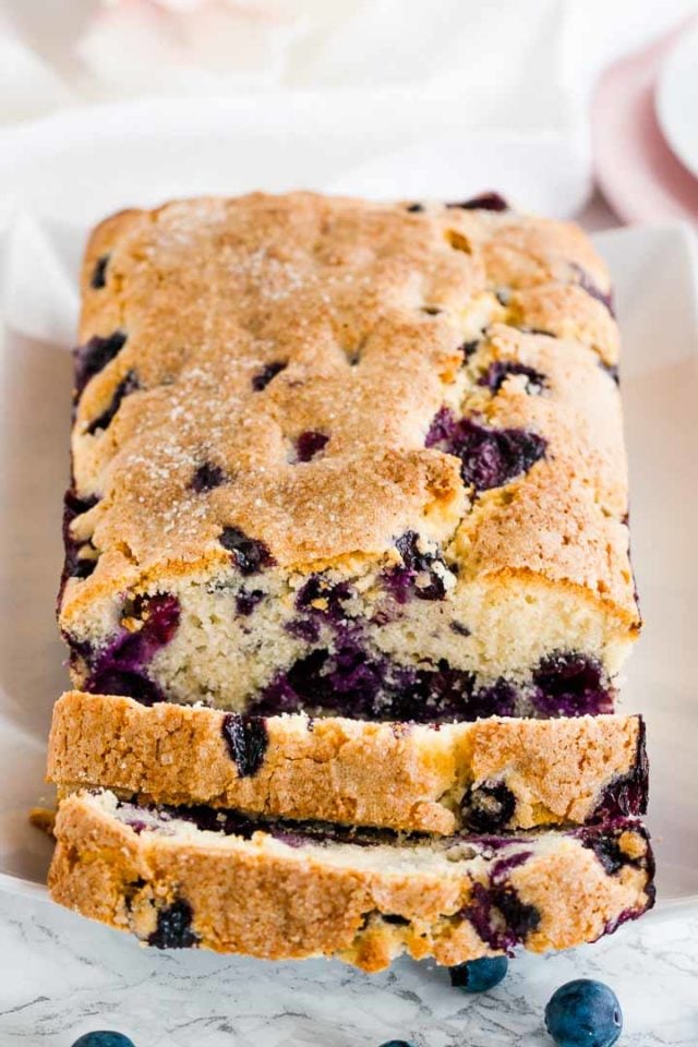 A loaf of blueberry bread on a white, rectangular plate with 2 slices cut off lying in front of it.