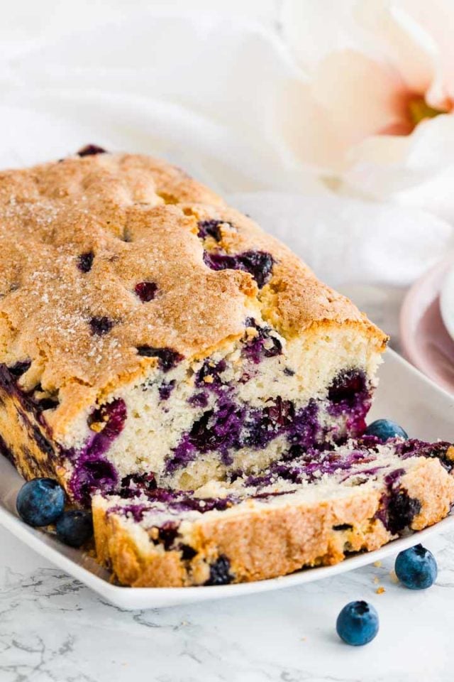 A loaf of blueberry bread on a white, rectangular plate with 1 slice cut off lying in front of it, garnished with blueberries and a flower.