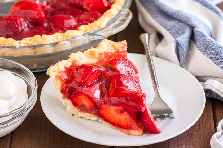 A slice of strawberry pie on a white plate with a silver fork on a wooden table. In the background, there\'s a glass baking dish with strawberry pie and a white and blue dish towel.