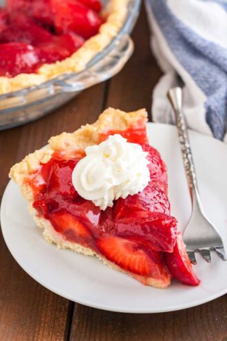 A slice of strawberry pie topped with whipped cream on a white plate with a silver fork on a wooden table. In the background, there's a glass baking dish with strawberry pie and a white and blue dish towel.