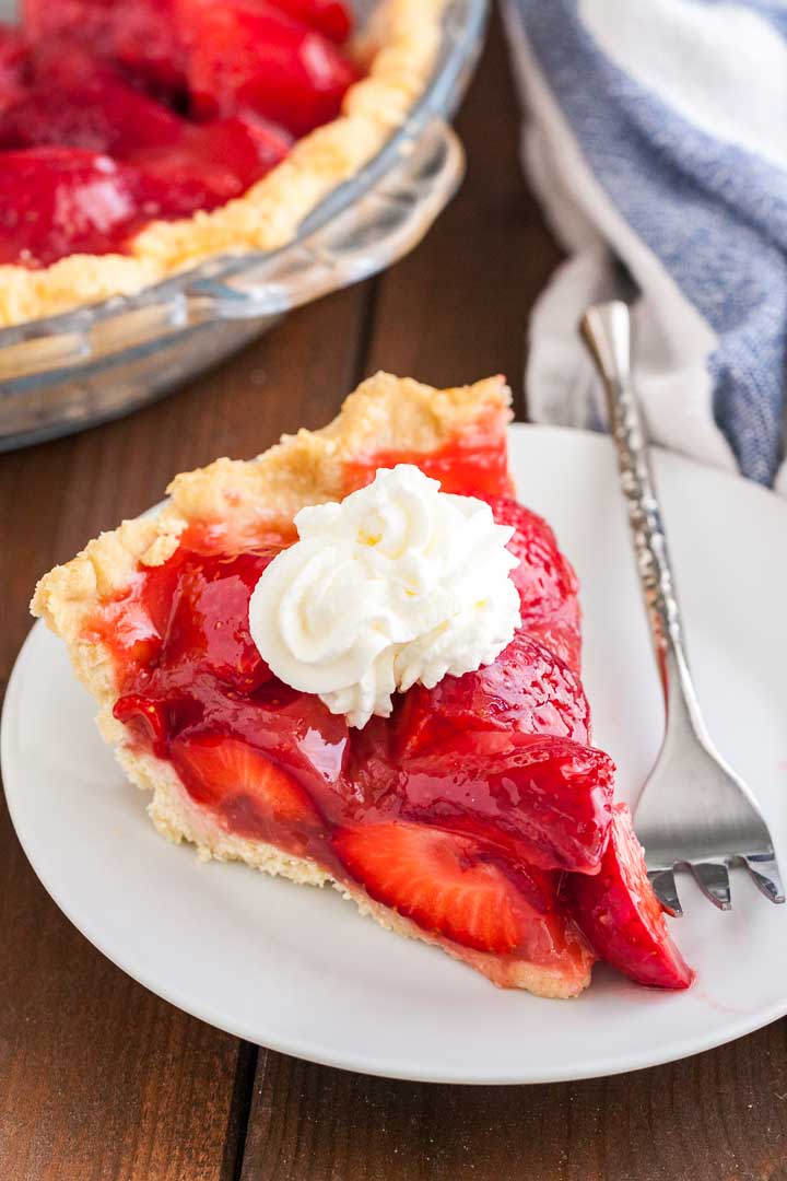 A slice of strawberry pie topped with whipped cream on a white plate with a silver fork on a wooden table. In the background, there\'s a glass baking dish with strawberry pie and a white and blue dish towel.