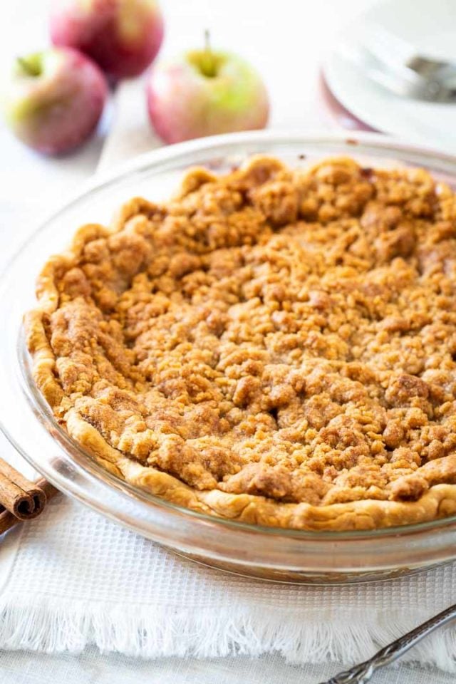 A glass baking dish, with apple pie with streusel topping garnished with apples and cinnamon sticks