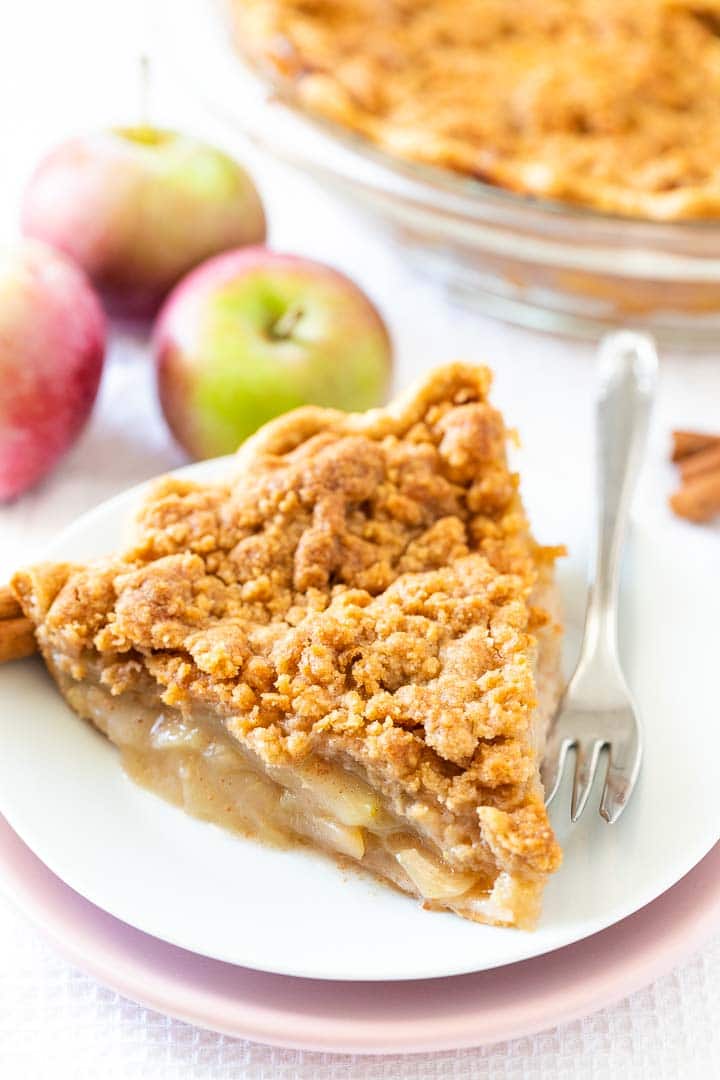 A piece of apple pie with streusel topping on a white plate stacked on a pink plate with a fork, topped with vanilla ice cream, garnished with cinnamon sticks, apples and a glass baking dish in the background.
