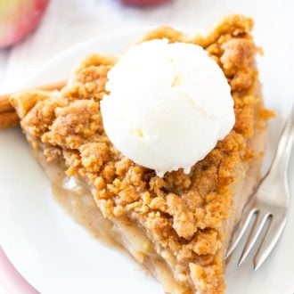 A piece of apple pie with streusel topping on a white plate stacked on a pink plate with a fork, topped with vanilla ice cream, garnished with cinnamon sticks and apple.