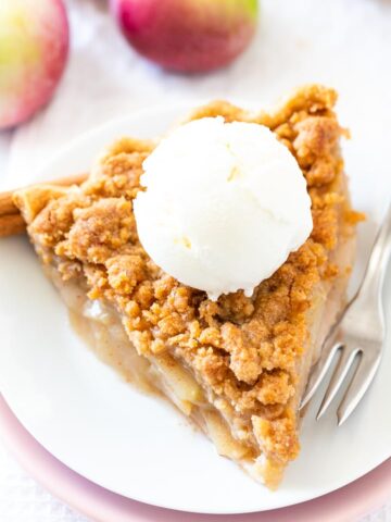 A piece of apple pie with streusel topping on a white plate stacked on a pink plate with a fork, topped with vanilla ice cream, garnished with cinnamon sticks and apple.