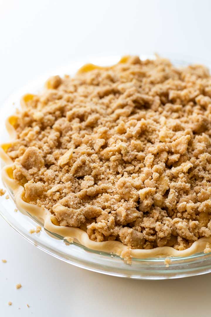 Close-up of glass baking dish with apple pie with streusel topping before baking. The crust is crimped into waves all around.