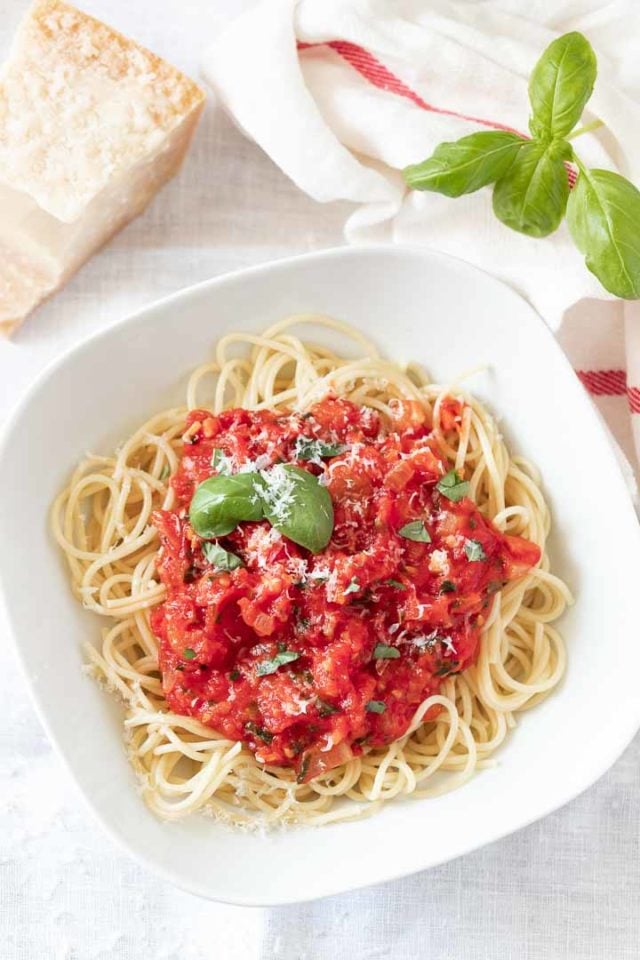 A white plate, with spaghetti topped with tomato sauce, garnished with basil leaves and parmigiano. Next to it, there\'s basil leaves, a white dish towel with a red stripe and a block of Parmigiano cheese.