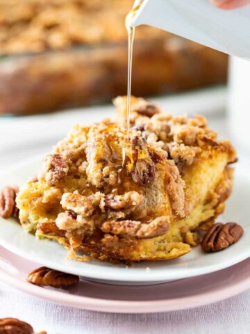 A piece of french toast casserole with pecans and streusel on a white plate stacked on a pink plate garnished with pecans. A small white jar is pouring maple syrup over it. There's a glass baking dish with the casserole in the background.