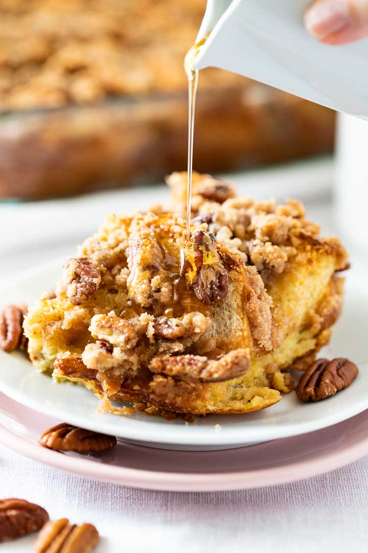 A piece of french toast casserole with pecans and streusel on a white plate stacked on a pink plate garnished with pecans. A small white jar is pouring maple syrup over it. There's a glass baking dish with the casserole in the background.