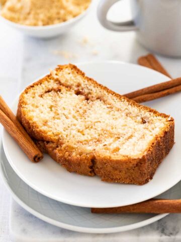 A white plate with a slice of cinnamon swirl bread, garnished with some cinnamon sticks and a grey canteen cup and a white bowl with cinnamon sugar mixture in the background.