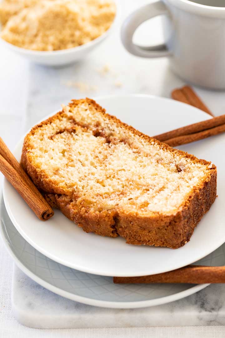 A white plate with a slice of cinnamon swirl bread, garnished with some cinnamon sticks and a grey canteen cup and a white bowl with cinnamon sugar mixture in the background.