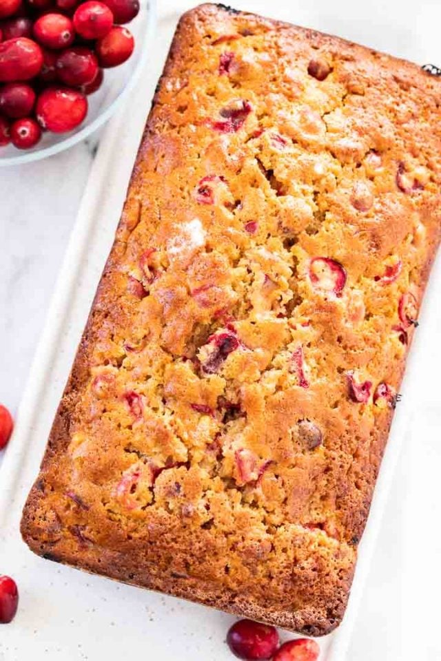 Top-down shot of cranberry orange bread on a rectangular porcelain serving platter next to a glass bowl of cranberries.