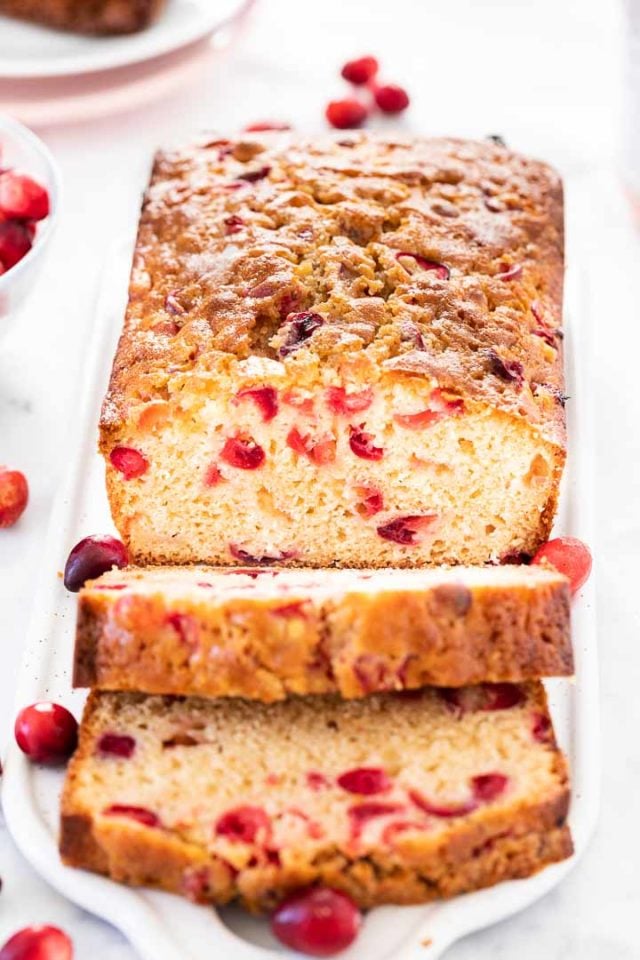 A Cranberry Orange Bread on a grey rectangular serving platter garnished with cranberries. Two slices have been cut off and are fallen over.