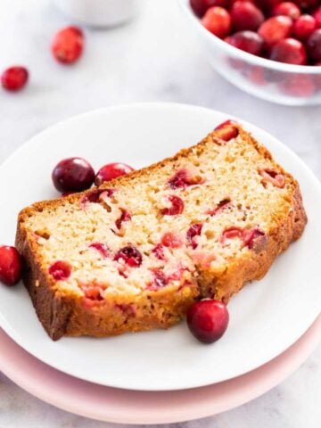 A slice of cranberry orange bread on a white plate stacked on a pink plate garnished with cranberries. There's a glass bowl with cranberries in the background.