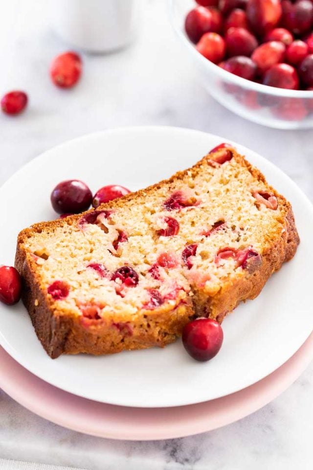 A slice of cranberry orange bread on a white plate stacked on a pink plate garnished with cranberries. There\'s a glass bowl with cranberries in the background.