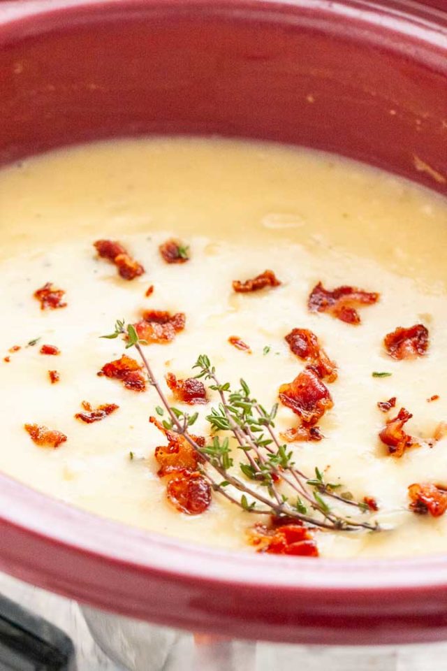 A red crockpot filled with potato soup topped with bacon crumbs and thyme.